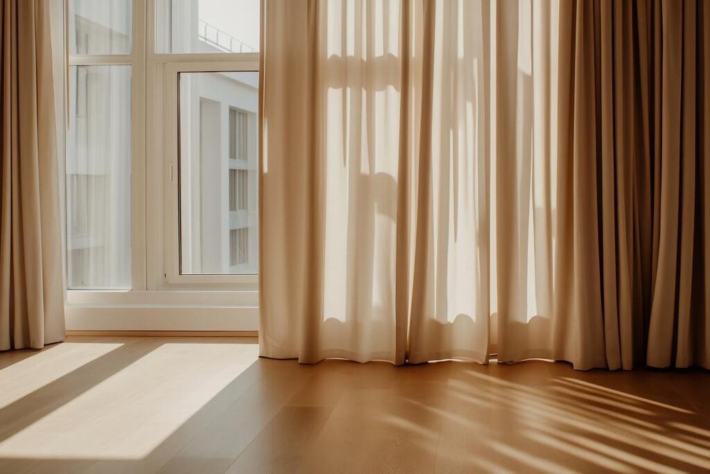 Clair Obscur Design, habillage de fenêtre, voilage beige , crème, baie vitrée, La lumière du soleil traverse les rideaux beiges, illuminant une pièce au sol recouvert de parquet.