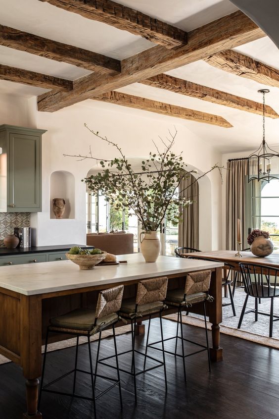 Clair Obscur Design, Interior Design, Airbnb Interior Design, Vacation Rental Decor, Short-Term Rental Design, modern organic kitchen and dining room, green steel shaker cabinets, central wooden island, quartz marble-effect countertop, wicker and wooden chairs, warm decor with natural branches and terracotta vases, ceiling beams, hanging chandelier, beige linen curtains, Italian style touches with arches and plaster walls, harmonious and eco-conscious design.